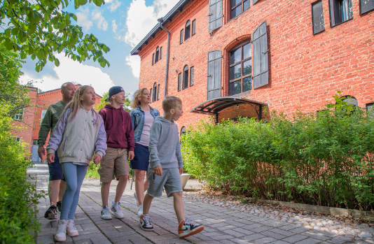 Perhe kävelyllä punatiilisten rakennusten edustalla kesäisissä vaatteissaan.