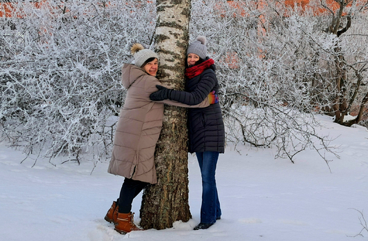 Kaksi naista halaamassa puuta talvisessa maisemassa.