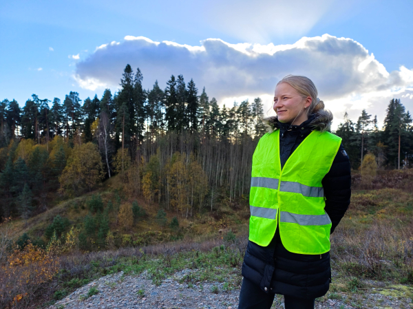 Nainen huomioliivipäällä seisomassa metsäisessä maisemassa aurinkoisena päivänä.