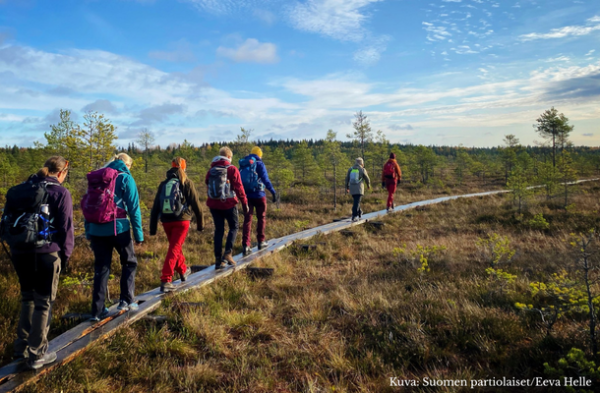 Ihmisiä retkeilemässä pitkospuilla suomaisemassa.