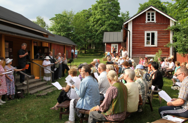 Ihmisiä yhteislaulutilaisuudessa Ronttismäen tehtaalaismuseon kesäisellä piha-alueella.